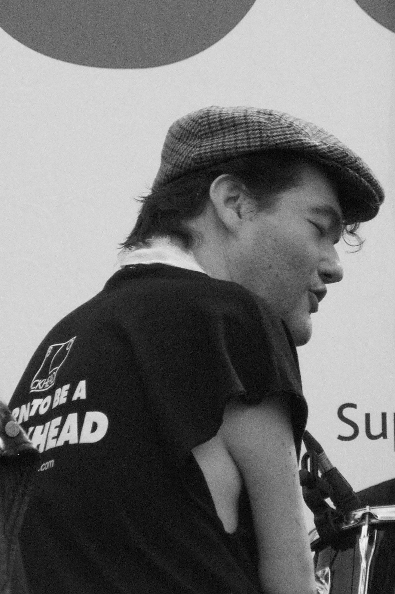 Black & white image: Back to camera, cap on with quiff, white scarf, sleeveless t-shirt with "Born To Be A Blockhead" written on the back